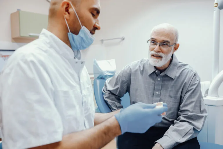A dental professional in a white coat, mask, and blue gloves holds a jaw model while consulting with an older adult wearing a gray shirt. The setting, which includes cabinetry and medical tools in the background, suggests a clinical environment focused on patient education or treatment planning.