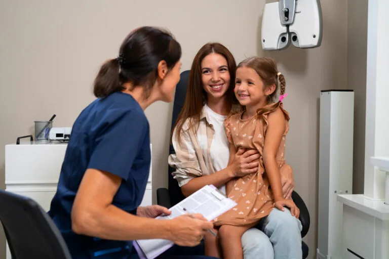 A healthcare professional in blue scrubs holds a document while speaking to a seated woman and a young girl on her lap, suggesting a family consultation. The room includes medical equipment like an eye exam device, indicating a clinical setting focused on patient education or routine check-ups.