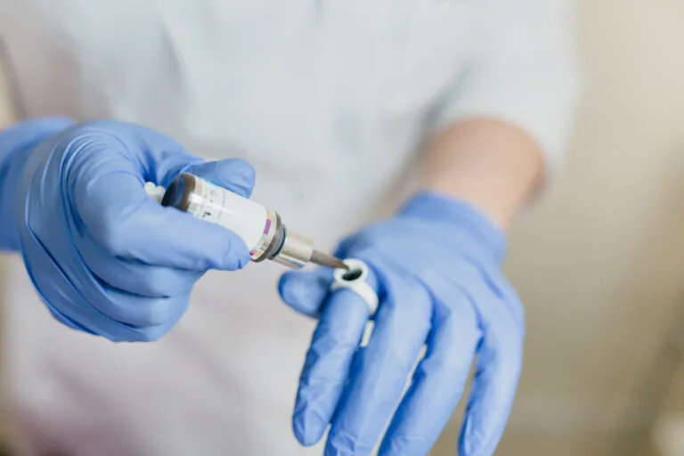 A gloved hand holds a syringe-like applicator angled toward a small vial, suggesting a precise injection or extraction procedure. The close-up composition and sterile setup highlight laboratory or medical practices where accuracy and safety are critical.